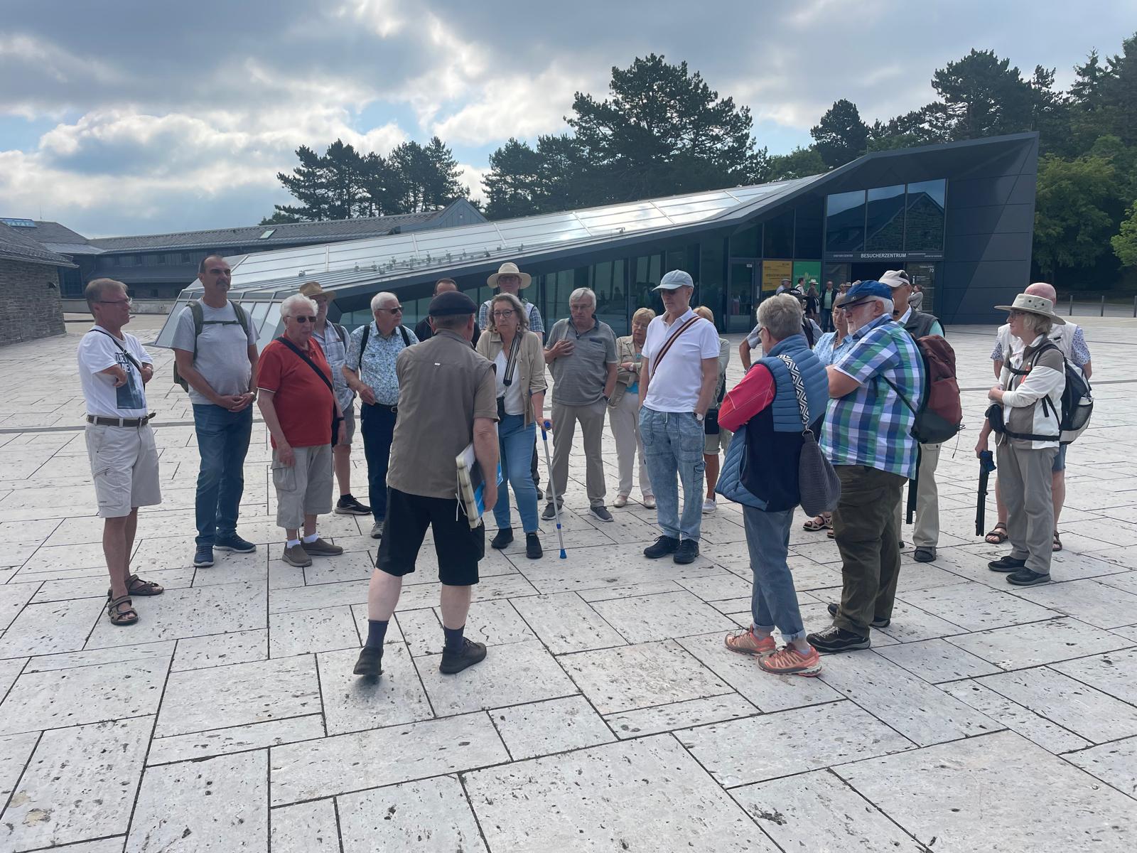 Eine Gruppe von Menschen steht auf einem gepflasterten Platz vor einem modernen Besucherzentrum und hört aufmerksam einem Reiseleiter zu.